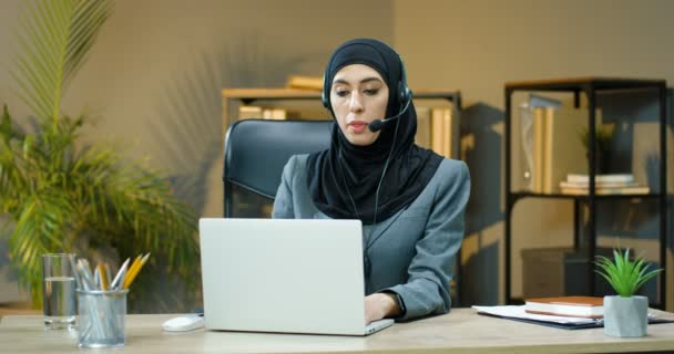 Retrato de mujer árabe joven en hijab y auriculares sentados en la mesa en la oficina, trabajando en el ordenador portátil y sonriendo a la cámara. Mujer musulmana mujer de negocios escribiendo en la computadora en el escritorio en el centro de llamadas. — Vídeos de Stock