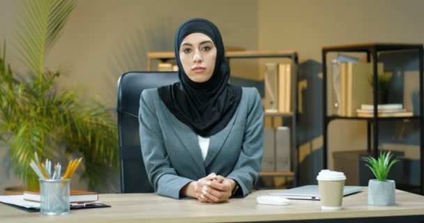 Retrato de la joven musulmana hermosa mujer en hijab negro sentado en la mesa en la oficina acogedora y sonriendo a la cámara felizmente. Bastante alegre mujer de negocios árabe en el escritorio en el gabinete con sonrisa. — Vídeos de Stock