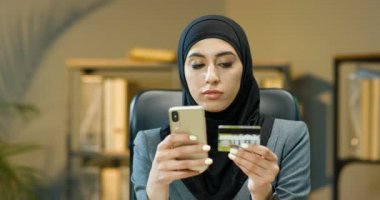 Geleneksel siyah tesettürlü oldukça mutlu genç Müslüman kadın akıllı telefondan alışveriş yapıyor ve ofiste otururken kredi kartıyla ödüyor. İnternetten telefon alan güzel bir Arap kadın..