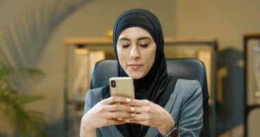 Siyah tesettürlü Arap güzeli genç bir kadın rahat bir ofiste masada oturuyor ve akıllı telefonları tıklatıyor ya da kaydırıyor. Geleneksel türban takmış Müslüman güzel iş kadını cep telefonundan mesaj atıyor