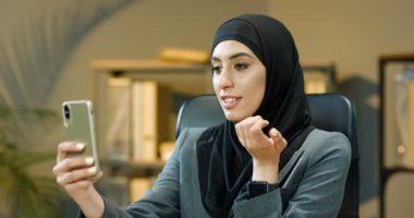 Geleneksel siyah tesettürlü neşeli müslüman kadın. Ofisteki müşterisiyle web kamerasıyla konuşuyor. Arap bayan yönetici telefonda video görüntülü sohbet ile müşteriyle konuşuyor.