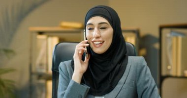 Geleneksel siyah başörtüsü takmış genç, güzel, neşeli bir Arap kadın cep telefonuyla konuşuyor ve rahat ofisinde gülümsüyor. Arap kadın cep telefonuyla sevinçle konuşuyor. Müslüman iş kadını arıyor..