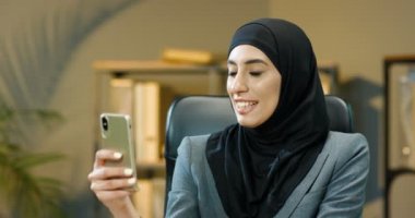 Geleneksel türbanlı genç, mutlu bir Arap kadın. Kabinedeki müşterisiyle internet kamerasıyla konuşuyor. Arap iş kadını telefonda video şapkalı müşteriyle konuşuyor. Video sohbeti.