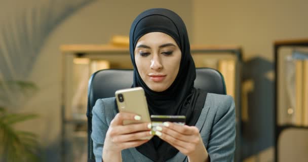 Jonge mooie vrolijke Arabische vrouw in traditionele zwarte hoofddoek winkelen online op de smartphone en betalen met een creditcard in het kantoor. Arabische vrouw kiezen en kopen in internet op mobiele telefoon. — Stockvideo