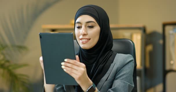 Mujer árabe bastante joven en hijab negro sentado en la oficina acogedora, pensando y tocando o desplazándose en la pantalla del dispositivo de la tableta. Muslim sonrió a la mujer de negocios en los mensajes de texto tradicionales pañuelo en la computadora. — Vídeo de stock