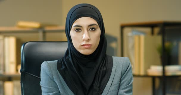 Πορτρέτο της νεαρής όμορφης μουσουλμάνα γυναίκα σε μαύρο χιτζάμπ κάθεται στο τραπέζι στο γραφείο και στροφή πρόσωπο με κάμερα. Γοητευτική σοβαρή Αραβίδα επιχειρηματίας με παραδοσιακή μαντίλα κοιτάζοντας ευθεία. — Αρχείο Βίντεο