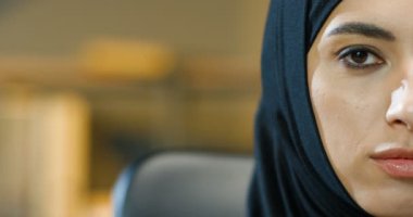 Kapalı mekanda siyah tesettürlü güzel müslüman kadının yarı kadın yüzünü kapatın. Ofisteki kameraya gülümseyen siyah gözlü Arap çekici kadının portresi. Geleneksel türbanlı iş kadını