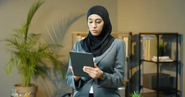 Siyah tesettürlü güzel Arap kadının portresi ofiste duruyor, tablet cihaz ekranını tıklatıyor ve kaydırıyor. Başörtülü Müslüman iş kadını kameraya gülümsüyor. Bilgisayar kullanan Arap kadın