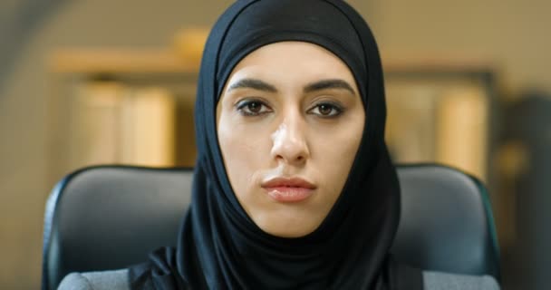 Primer plano de la cara femenina de la hermosa joven musulmana en el interior de hijab negro. Retrato de la encantadora dama árabe con los ojos oscuros sonriendo a la cámara en la oficina. Mujer de negocios en pañuelo para la cabeza tradicional. — Vídeos de Stock