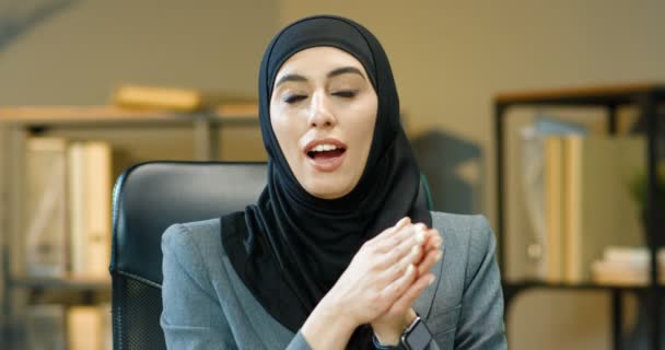 Joven mujer musulmana hermosa en hijab negro sentado en la mesa en la oficina y hablando con la cámara mientras educa. Bonito bloguero de negocios árabe encantador o cauch en el tradicional pañuelo que habla. — Vídeos de Stock