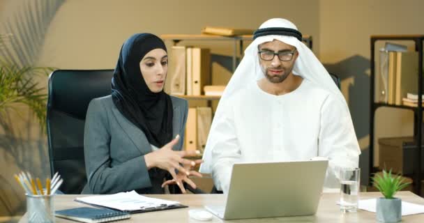 Hombres y mujeres árabes, colegas de negocios hablando y discutiendo el proyecto en la computadora portátil en la oficina. Una mujer en Hijab y un hombre en Kandura de Emiratos Árabes Unidos trabajando en la computadora. Empresario y empresaria en emiratos — Vídeos de Stock