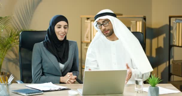 Muslimische Kollegen sitzen am Tisch im Büro, reden und arbeiten am Computer in den Vereinigten Arabischen Emiraten. Arabische schöne Geschäftsfrau im Hijab und Geschäftsfrau im Kandura, Kollegen diskutieren über die Arbeit am Laptop. — Stockvideo