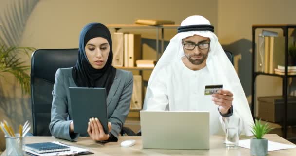 Emirates hombre de negocios en kandura compras en línea en el ordenador portátil y el pago con tarjeta de crédito en la oficina. Asistente femenina en hijab ayudando con el dispositivo de tableta árabe musulmán hombre y mujer que trabajan en el escritorio — Vídeos de Stock