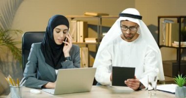 Arap genç iş kadını tesettürlü cep telefonuyla konuşuyor ve ofiste dizüstü bilgisayarla çalışıyor. Kandura 'daki Arap işadamı tablet cihazını tıklatıyor ve kaydırıyor. BAE 'den birkaç meslektaşım..