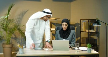 Siyah başörtüsü takan Müslüman kadın ofiste oturuyor ve dizüstü bilgisayarla çalışıyor. Bulaşıkçı Adam yanında duruyor ve asistan projesini kontrol ediyor. Erkek yardım ve danışmanlık.