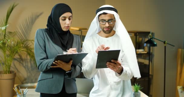 Muslimský podnikatel v kanduře a brýle klepání a rolování na tablet zařízení a ženské asistentky poznámku v dokumentu ve složce. Arabský muž a žena v tradičních emirátech oblečení pracující v úřadu — Stock video