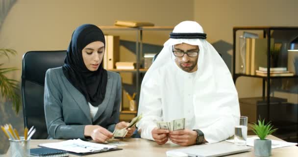Muzułmański bogaty biznesmen i bizneswoman siedzi przy biurku w biurze i pracuje. Kobieta w hidżabie i mężczyzna w Kandurze liczący pieniądze i dolary. Arabski samiec i samica pisząca ilość banknotów. — Wideo stockowe