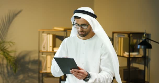 Arabo bell'uomo in occhiali e Kandura in piedi in ufficio, toccando e scorrendo sullo schermo del dispositivo tablet. Uomo d'affari musulmano in abiti tradizionali emirates. Maschio arabo che usa il computer. Affari degli Emirati Arabi Uniti — Video Stock