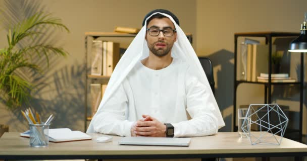 Joven hombre musulmán guapo en Kandura sentado en la mesa en la oficina y hablando con la cámara mientras educa. Blogger árabe de negocios o cauch en emiratos tradicionales manswear hablando. Hombre de Emiratos Árabes Unidos. — Vídeos de Stock