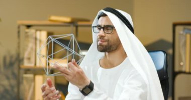 Beyaz Kandura 'lı yakışıklı genç bir Arap' ın portresi rahat ofisindeki masada oturan, düşünen ve geometrik figürü tutan gözlüklü. Müslüman erkek işadamı iş meselelerini düşünüyor.