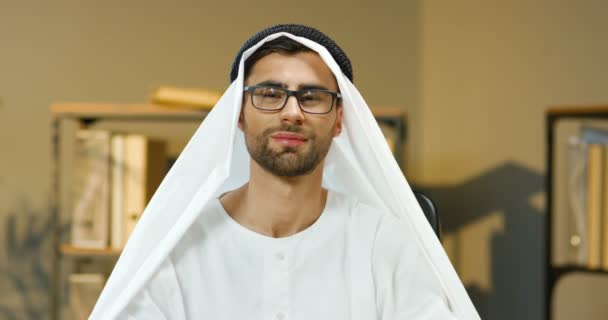 Portraitaufnahme eines jungen gut aussehenden muslimischen Mannes in Diadem, der im Büro am Tisch sitzt und in die Kamera lächelt. Arabischer Geschäftsmann im traditionellen Emirat-Outfit blickt mit einem Lächeln geradeaus. — Stockvideo