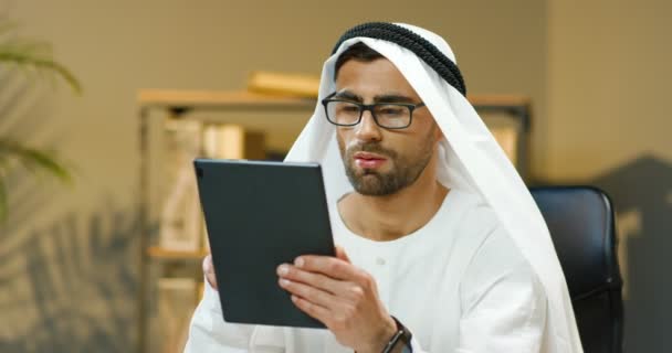 Arabisch gut aussehender junger Mann in Kandura sitzt am Tisch im gemütlichen Büro und tippt oder blättert auf dem Tablet-Gerät. Der muslimische Geschäftsmann schreibt SMS und chattet auf einem Gadget-Computer. Arabische Männchen. — Stockvideo