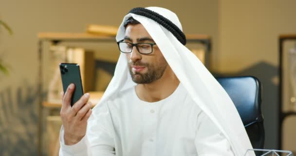 Красивый мусульманский молодой человек в посудомойке сидит за столом в офисе и разговаривает через веб-камеру по мобильному телефону. Арабский бизнесмен в Кандуре ведет видеочат на смартфоне. Видеочат на мобильном телефоне. — стоковое видео