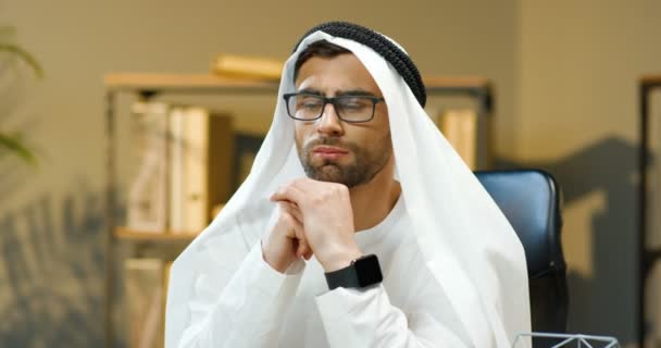 Retrato de un joven hombre musulmán guapo en Kandura y gafas sentadas en la oficina y considerando algo. Empresario árabe en traje de emiratos tradicionales pensando en los negocios. Hombre en dishdasha. — Vídeos de Stock