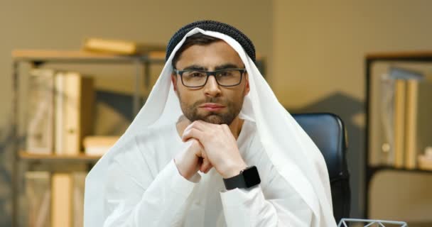 Retrato de un joven hombre musulmán guapo en Kandura y gafas sentadas en la oficina y sonriendo a la cámara. Hombre de negocios árabe en traje de emiratos tradicionales mirando directamente con sonrisa. Hombre en dishdasha. — Vídeos de Stock