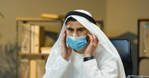 Portrait of young handsome muslim man in Kandura taking off medical mask and smiling to camera in office. Arabic businessman in traditional emirates outfit with smile. Male in dishdasha. Pandemic time — Stock Video