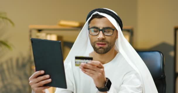 Молодой мусульманин в традиционных эмиратах покупает одежду онлайн на планшете и платит кредитной картой, сидя за столом в офисе. Красивый арабский мужчина покупает в интернете на гаджете в ОАЭ. — стоковое видео