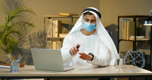Arabczyk w Kandurze i maska medyczna siedzący przy biurku, dezynfekujący ręce środkiem dezynfekującym i pracujący na laptopie. Męski muzułmanin czyści skórę środkiem dezynfekującym. Arabski przedsiębiorca. — Wideo stockowe