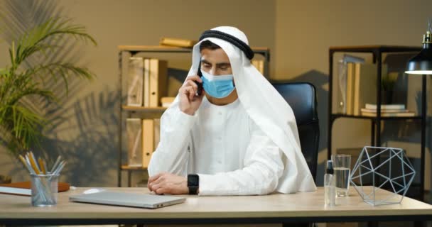 Homem árabe em máscara médica e dishdasha sentado na mesa no escritório, falando no celular e trabalhando. Muslim masculino na roupa emirates tradicional que fala no telefone. Árabe empresário bem sucedido . — Vídeo de Stock