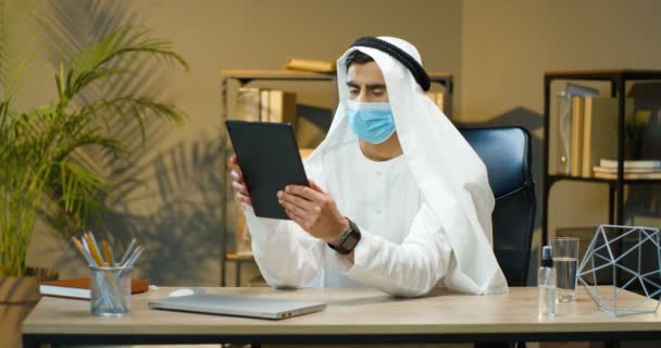 Hombre joven guapo árabe en Kandura y máscara médica sentado en la mesa en la oficina acogedora y golpeando o desplazándose en el dispositivo de la tableta. Hombre de negocios musulmán mensajes de texto y chat en la computadora gadget. — Vídeos de Stock