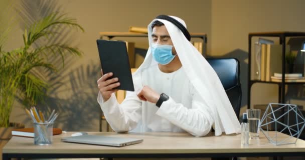 Przystojny muzułmanin w dishdashy i maska medyczna siedzący przy stole w biurze i rozmawiający przez kamerę internetową na tablecie. Arabski biznesmen w Kandurze ma video z gadżetem. Wideokatowanie. — Wideo stockowe