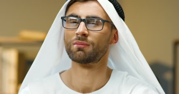 Крупным планом мужского лица красивого молодого мусульманина в белом помещении Кандуры. Портрет арабского парня с темными глазами в очках, улыбающегося в камеру в офисе. Бизнесмен в традиционной одежде эмиратов. — стоковое видео