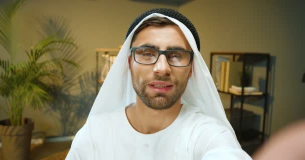 POV молодого красивого мусульманина в белом традиционном эмирате эмигрирует Кандура и очки разговаривают с камерой во время видеочата в офисе. Арабский мужчина в видеочате на дишдаше через веб-камеру в помещении. — стоковое видео