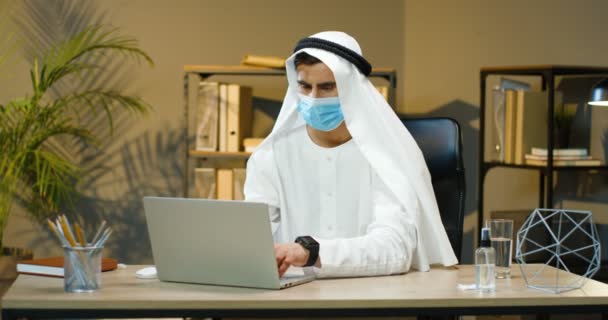 Arabczyk w Kandurze i maska medyczna siedząca przy biurku i pracująca na laptopie. Mężczyzna muzułmanin menedżer w tradycyjnych emiratów ubrania pisząc na komputerze i uśmiechając się do kamery. Arabski przedsiębiorca. — Wideo stockowe