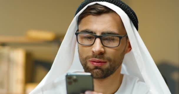 Közelkép egy jóképű fiatal muszlim férfiról fehér kandúrában, aki a mobilján tapogatózik és teker. Arab fickó szemüvegben sms-t ír okostelefonon az irodában. Üzletember hagyományos EAE ruhákban. — Stock videók