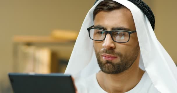 Крупным планом красивого молодого мусульманина в белой кандуре и очках, стучащих и прокручивающихся по планшетному устройству. Арабский парень в очках пишет смс или просматривает гаджет в офисе. Бизнесмен в ОАЭ. — стоковое видео