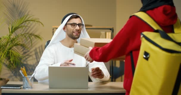 Arabiska ung stilig man i Kandura och glasögon sitter vid bord i kontor och arbetar. Kurir levererar lådor. Leverantör föra paket lådor till muslimsk affärsman i skåp. — Stockvideo
