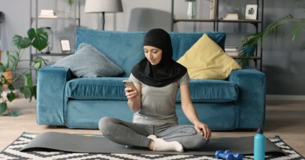 Jonge Arabische vrouw in traditionele hoofddoek zittend op mat op de vloer in sportieve kleding en sms-bericht op smartphone. Thuis. Arabische vrouw in hijab glimlachen tijdens het afluisteren en scrollen op de telefoon. — Stockvideo