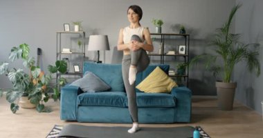 Gülen esmer, beyaz tenli, spor elbiseli güzel bir kadın. Salonda bacakları ve kalçaları esnetiyordu. Güzel bir kadın yoga yapıyor ve evde egzersiz yapıyor. Dengeleme ve egzersiz.