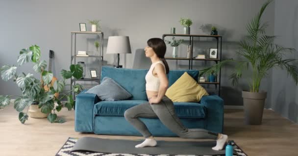 Young beautiful slim Caucasian woman squatting in cozy living room and exercising for strong legs in shape. Pretty female doing squats at home. Crouches for hips and butt in shape. — Wideo stockowe
