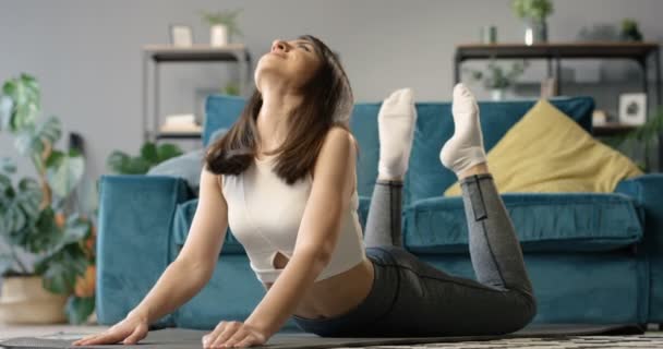 Bela jovem caucasiana esportiva mulher esticando as costas e curvando-se sobre tapete de ioga no chão em casa. Muito feminino yogi exercitando e fazendo barco asana pose na sala de estar aconchegante. — Vídeo de Stock