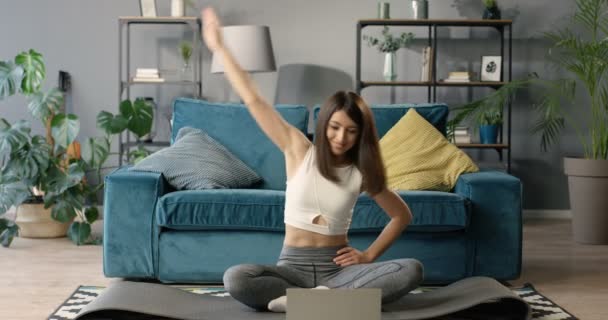 Pretty young Caucasian woman sitting on yoga mat on floor in front of the laptop computer at home. Attractive slim girl working out indoor with internet video course. Lesson with instructor online. — Stock Video