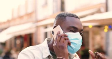 Tıbbi maskeli genç Afrikalı Amerikalı adam sokakta yürüyor ve cep telefonuyla konuşuyor. Yakışıklı bir erkek cep telefonuyla konuşuyor ve geziniyor. Açık havada. Telefon görüşmesi.