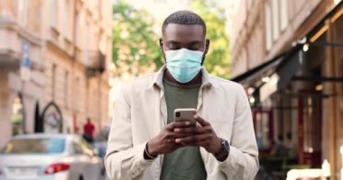 Tıbbi maskeli genç Afrikalı Amerikalı adam şehir caddesinde yürüyor ve akıllı telefondan mesaj yolluyor. Yakışıklı erkek dinleme ve kaydırma cep telefonu ve gezinti. Açık hava..