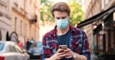 Tıbbi maskeli genç beyaz adam sokakta yürüyor ve akıllı telefondan mesaj yolluyor. Yakışıklı bir erkek cep telefonuna takılıp kayıyor, şehirde kayıyor. Dışarıda..