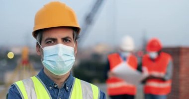 Beyaz tenli, orta yaşlı, yakışıklı bir inşaatçının portresi. İnşaatta durup kameraya bakıyor. Miğferli erkek müteahhitin etrafını sarın. Salgın.